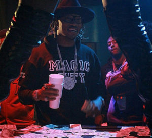Rapper Future wearing a Magic City strip club hoodie and a large diamond chain holding a double cup and throwing money at a stripper