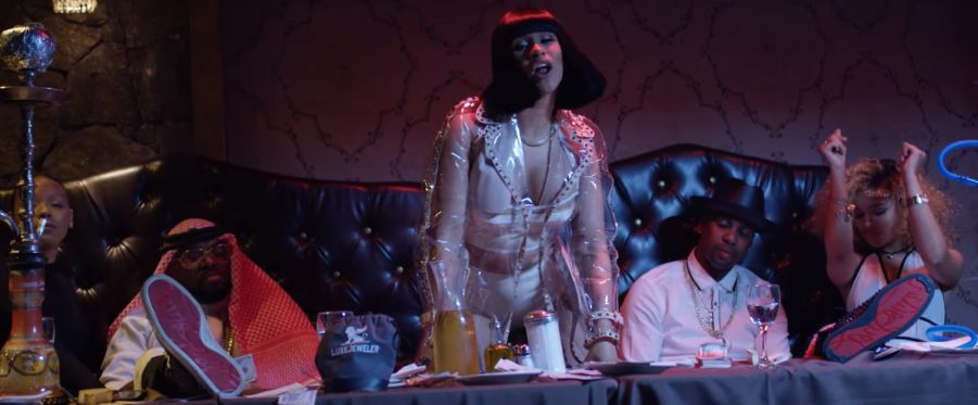 Cardi B in Bodak Yellow video. Four people sit on a black leather couch behind her. Two men are wearing Christian Louboutin shoes.