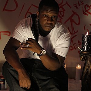 Hip hop artist Picasso De'France on set of music video for Running, sitting in front of painted wall with angels. He is wearing a white shirt with a black bandana over his shoulder. Wearing a watch and diamond ring.